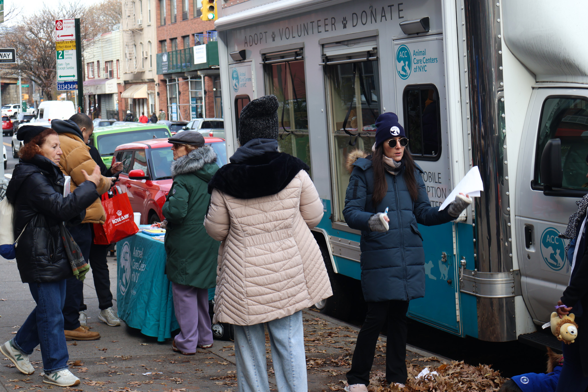 An Animal Care Centers of NYC volunteer in a branded beanie handing paperwork to a visitor beside the mobile adoption truck