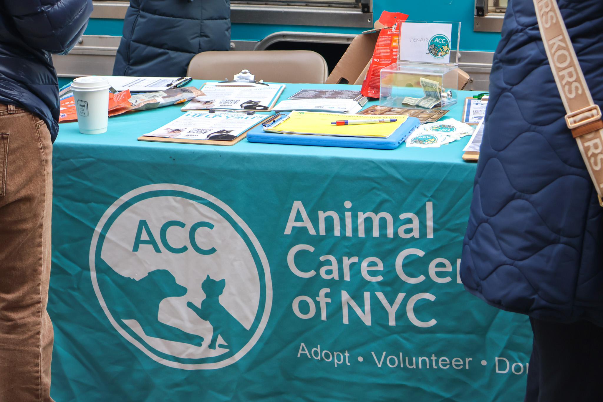 An Animal Care Centers of NYC table covered in clipboards, pamphlets, and a clear donation box, seen from behind two visitors