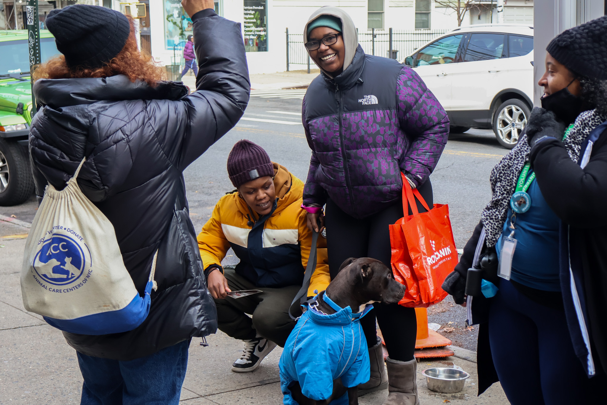 A group of smiling visitors gathered around a newly adopted dog in a blue coat, one of them carrying an Animal Care Centers of NYC tote bag