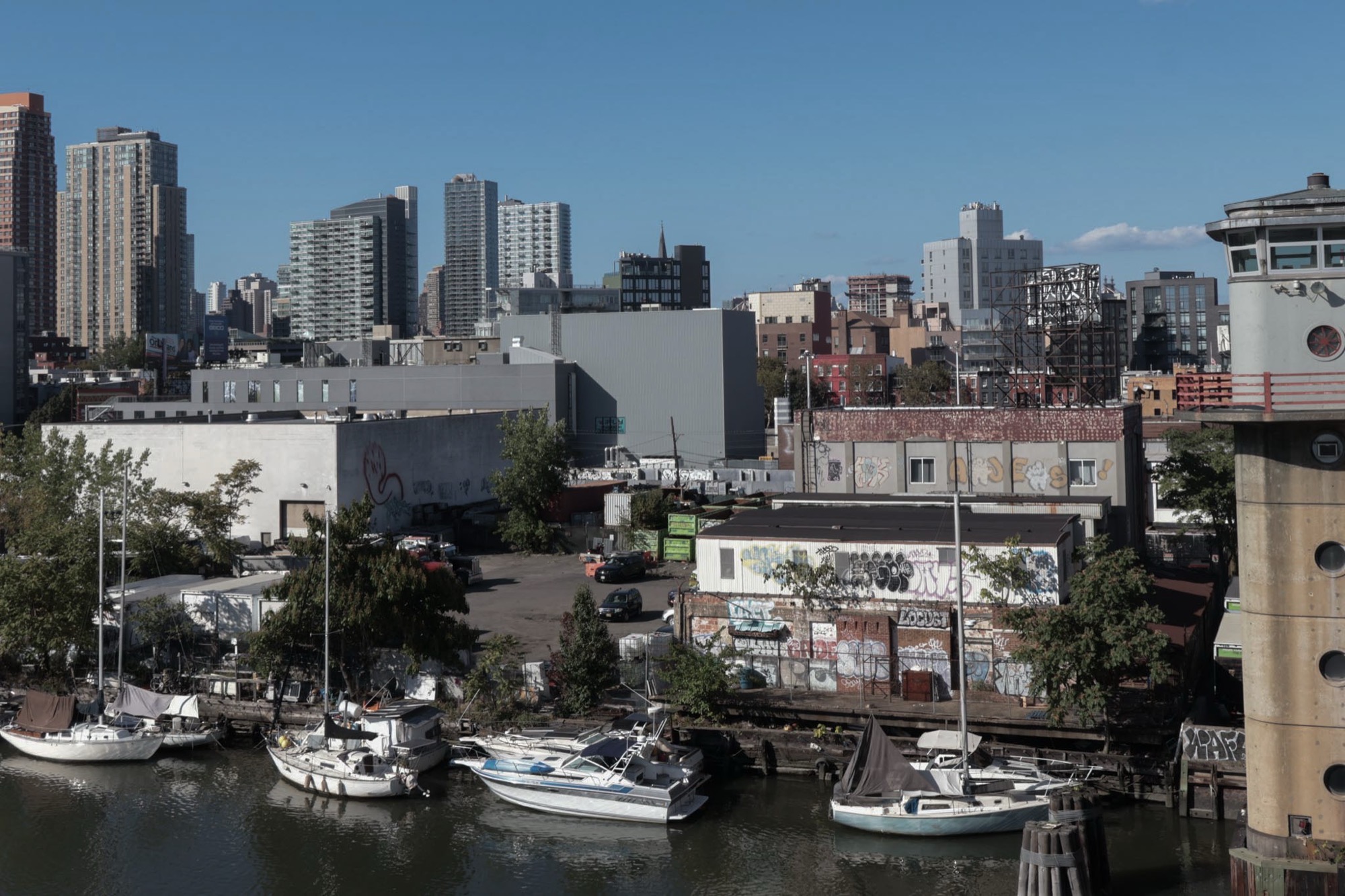 A calm marina on Newtown Creek with sailboats, brick buildings, and the Manhattan skyline in the distance