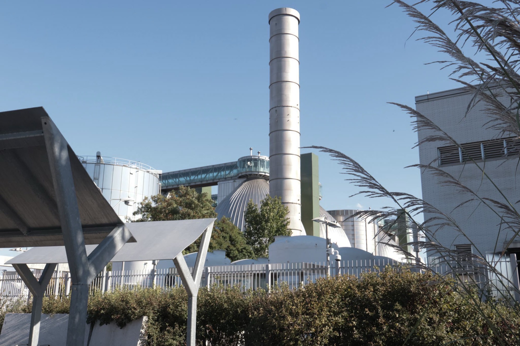 A wastewater treatment stack rising behind a chain-link fence and shrubs