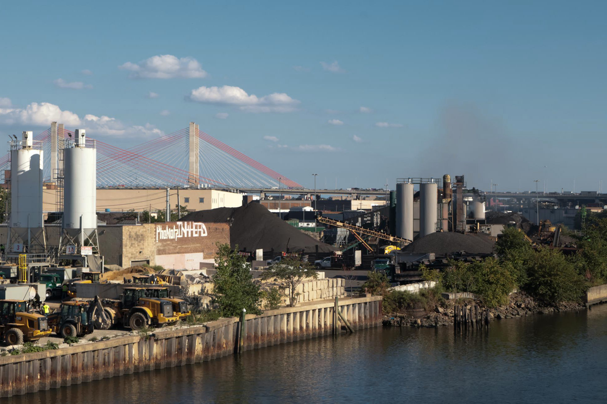 An industrial waterfront on Newtown Creek with smokestacks and a plume of pink-tinted smoke against a blue sky