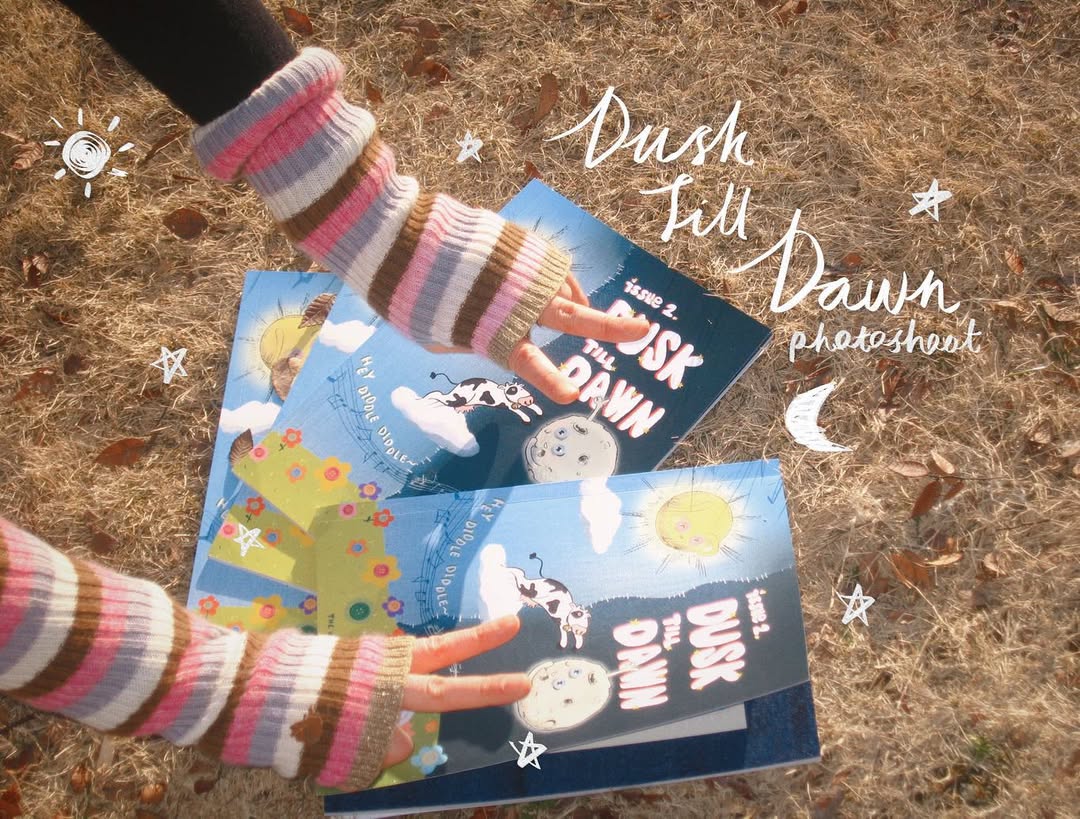 Overhead shot of hands holding two copies of Dusk Till Dawn on dry grass, with 'Dusk Till Dawn photoshoot' hand-lettered over the photo